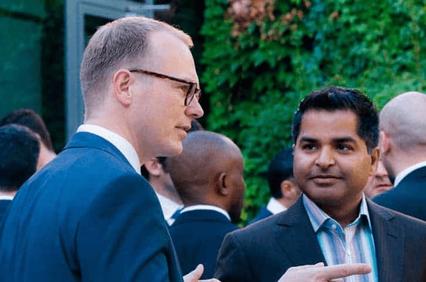 Two men conversing at an outdoor event, one pointing gently while discussing.